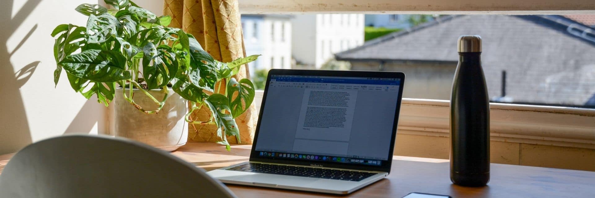 A home desk facing an open window, letting in the sun. The desk has an open laptop, bottle of water, mobile phone and a green houseplant on it.